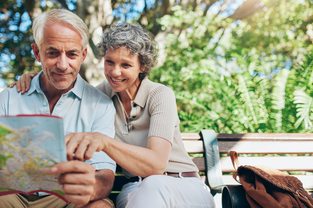 A senior man and a senior woman sit on a park bench and look at a paper map