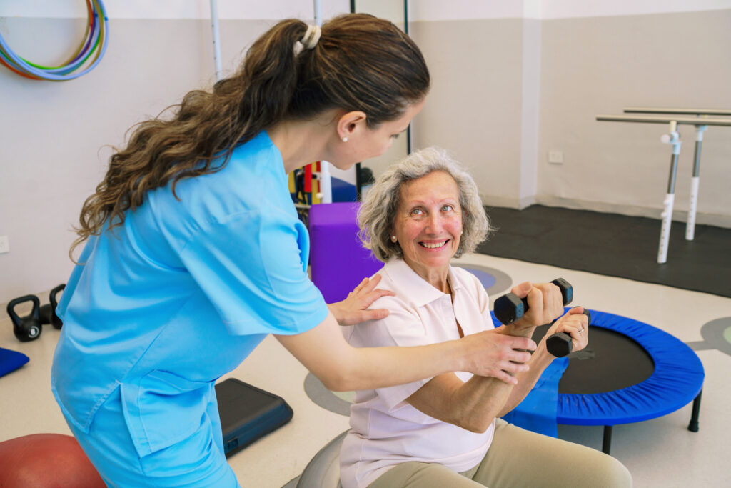 A friendly physiotherapist guides a senior woman through dumbbell exercises in a rehabilitation clinic, promoting fitness, mobility, and strength training for healthy aging.
